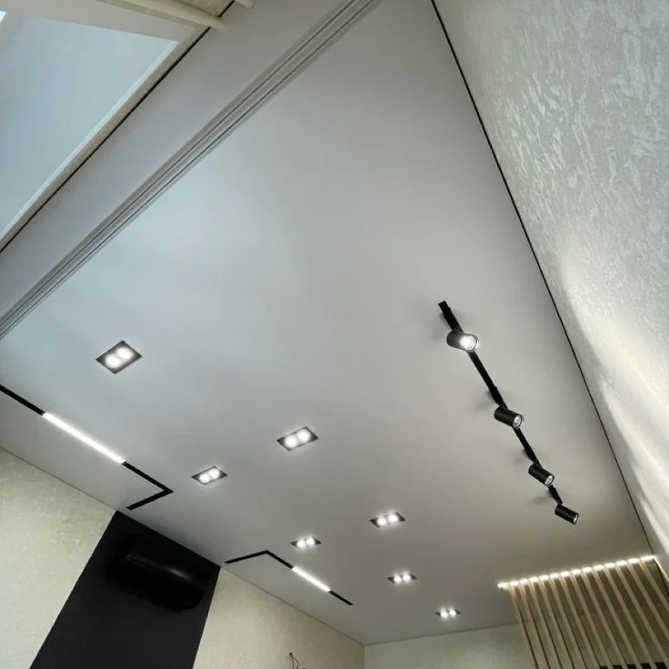Nowoczesny, biały sufit z oświetleniem LED: punktowe reflektory na szynie i wpuszczane kwadratowe lampy. Dekoracyjne wykończenie ścian w jasnych odcieniach.