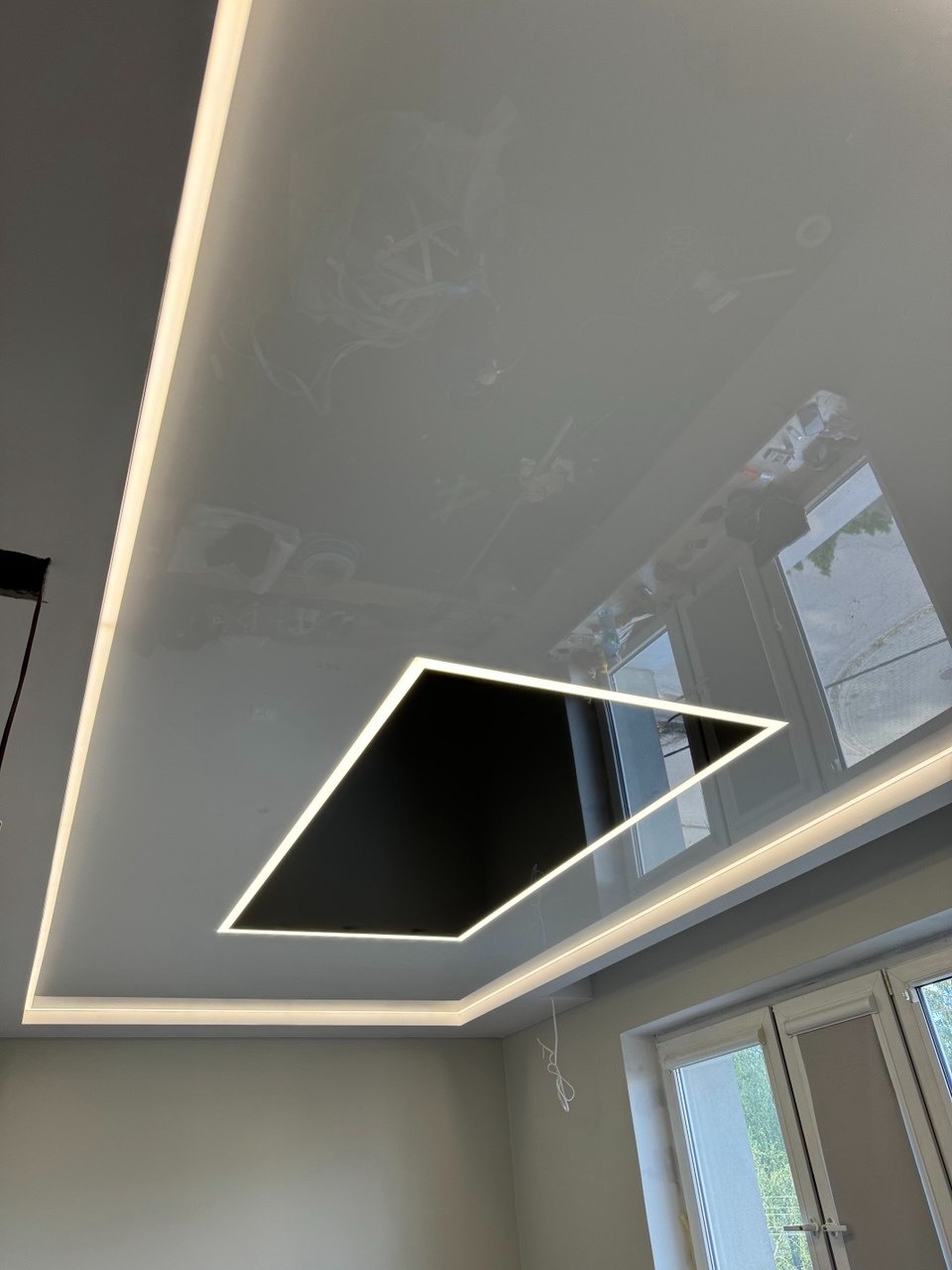 Błyszczący, biały sufit z czarnym, wpuszczonym elementem w kształcie rombu, obwiedzionym ledowym oświetleniem. Widoczne odbicie okna.