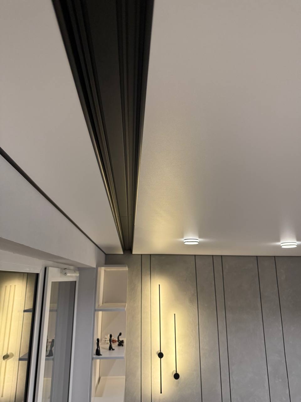 Nowoczesny sufit z czarną listwą i oświetleniem punktowym. Ściana z dekoracyjnymi lampami liniowymi. Minimalistyczne wnętrze z elementami wykończenia.