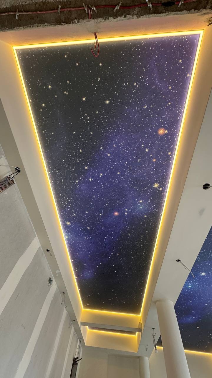 Nietypowy sufit z motywem kosmosu, podświetlony ciepłym światłem LED, tworzący unikalny efekt wizualny. Widoczne niedokończone ściany w tle.