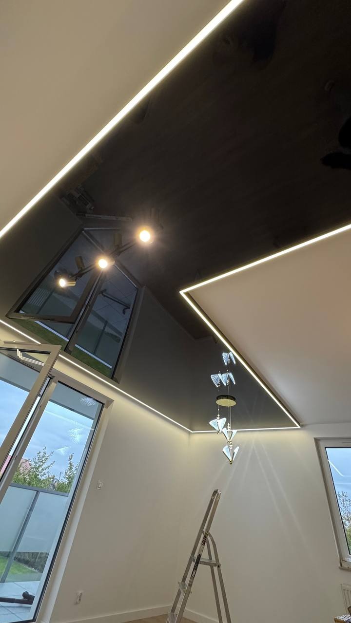 Nowoczesny, czarny sufit z oświetleniem LED wzdłuż krawędzi, nietypowe okno dachowe i designerska lampa wisząca. Widok z dołu, drabina w rogu pokoju.