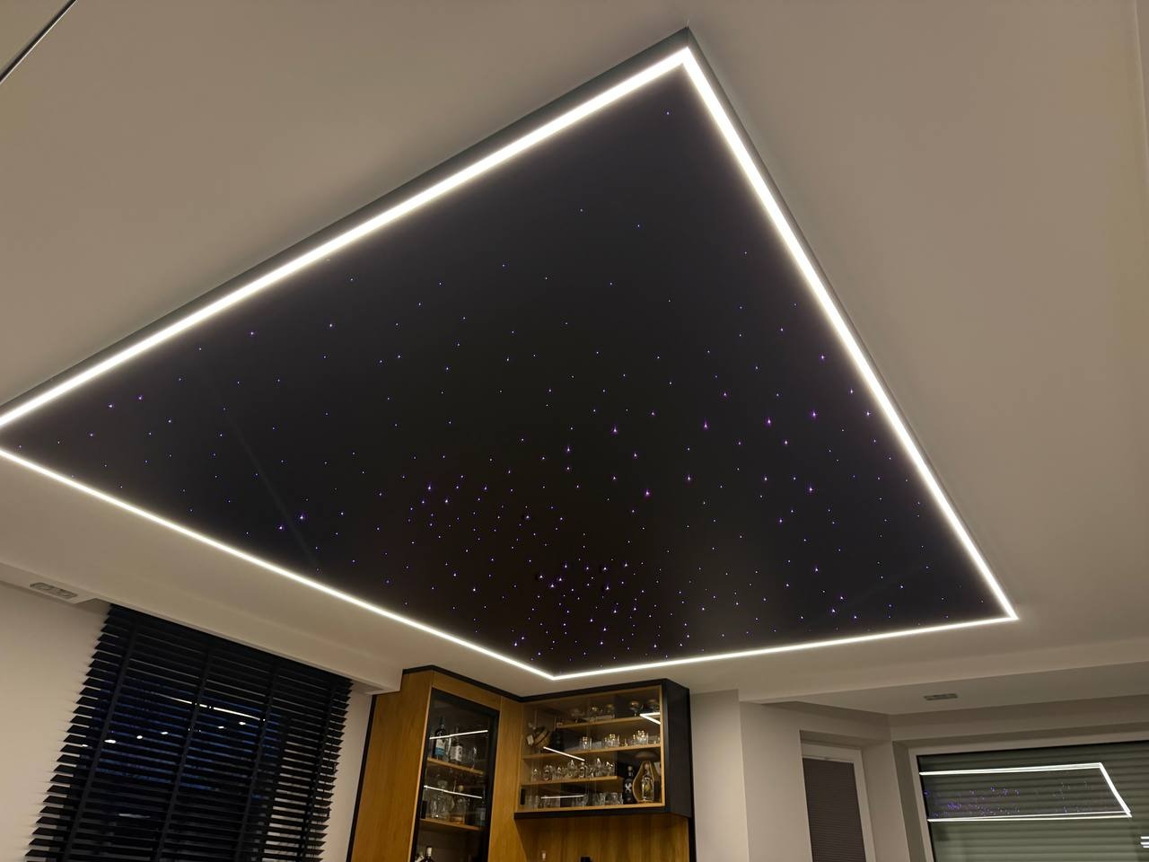 Nowoczesny sufit z efektem gwieździstego nieba, obramowany listwami LED. Widok na część umeblowanego wnętrza z żaluzjami i przeszkloną witryną.