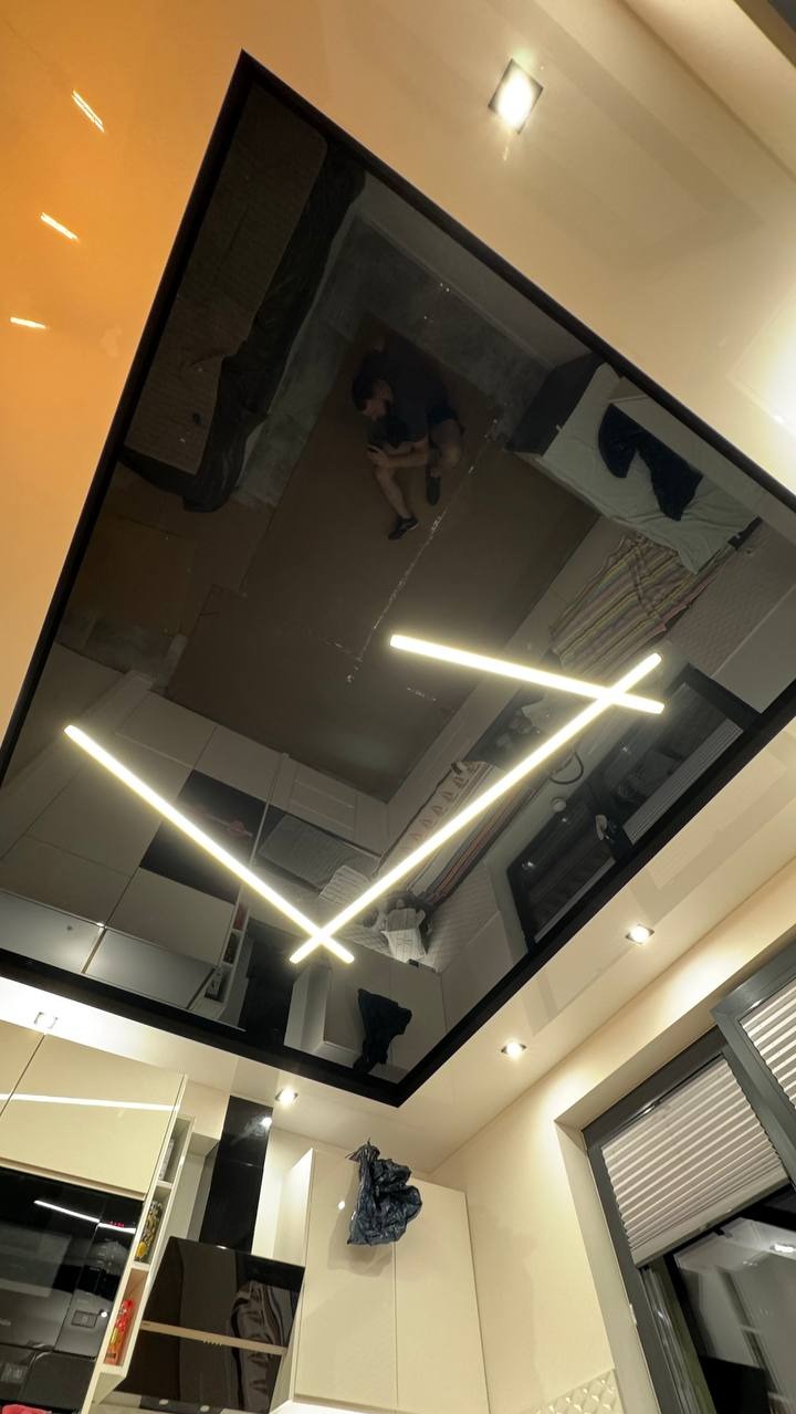 Błyszcząca, czarna powierzchnia sufitu z wbudowanym oświetleniem LED w kształcie krzyża, odbijająca wnętrze pokoju. Widoczny fragment kuchni i okno z żaluzjami.