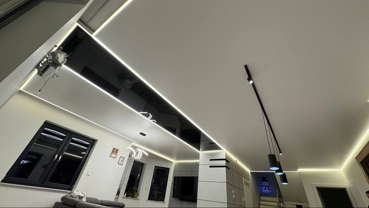 Nowoczesny salon z czarnym, połyskującym sufitem i liniowym oświetleniem LED. Widoczne okna, lampy wiszące i elementy dekoracyjne. Minimalistyczny design.