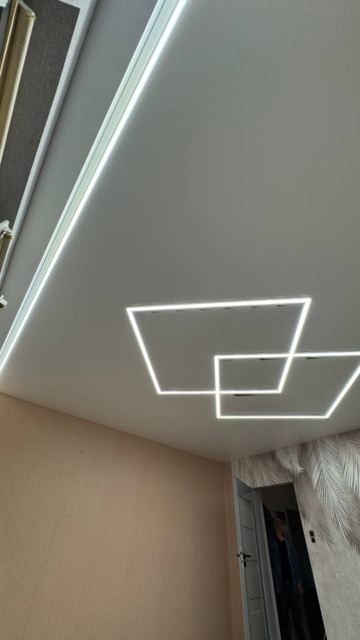 Biały sufit z podwieszanymi, kwadratowymi lampami LED, tworzącymi geometryczny wzór. Widoczny fragment ściany i drzwi z wzorzystą tapetą.