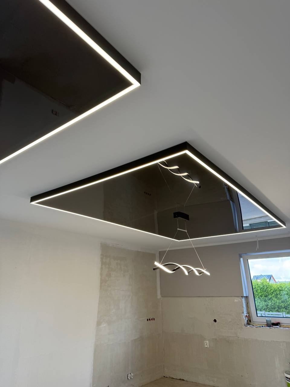 Czarny, lustrzany sufit napinany z oświetleniem LED w kształcie prostokąta, nowoczesna lampa wisząca, ściany w trakcie wykończenia.