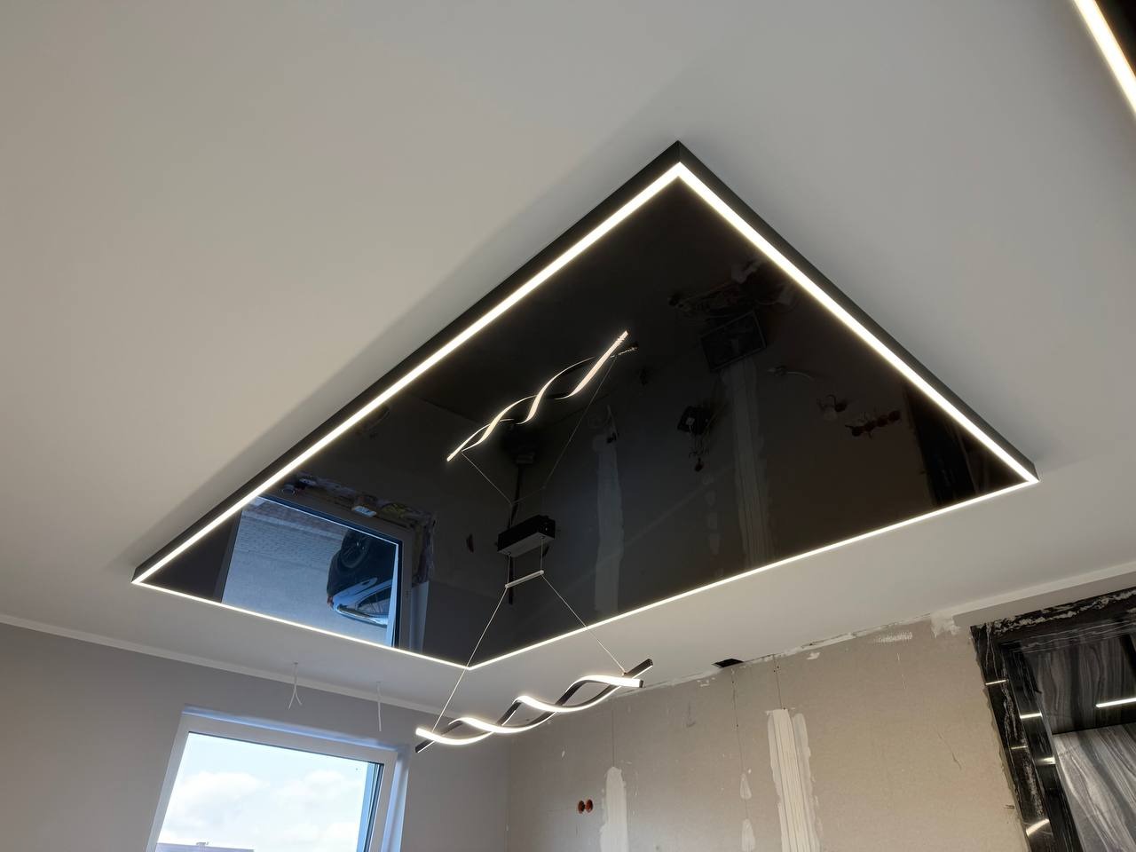 Błyszczący, czarny sufit z oświetleniem LED w kształcie prostokąta, odbijający okno i wiszącą lampę. Widoczne ściany w trakcie wykończenia.