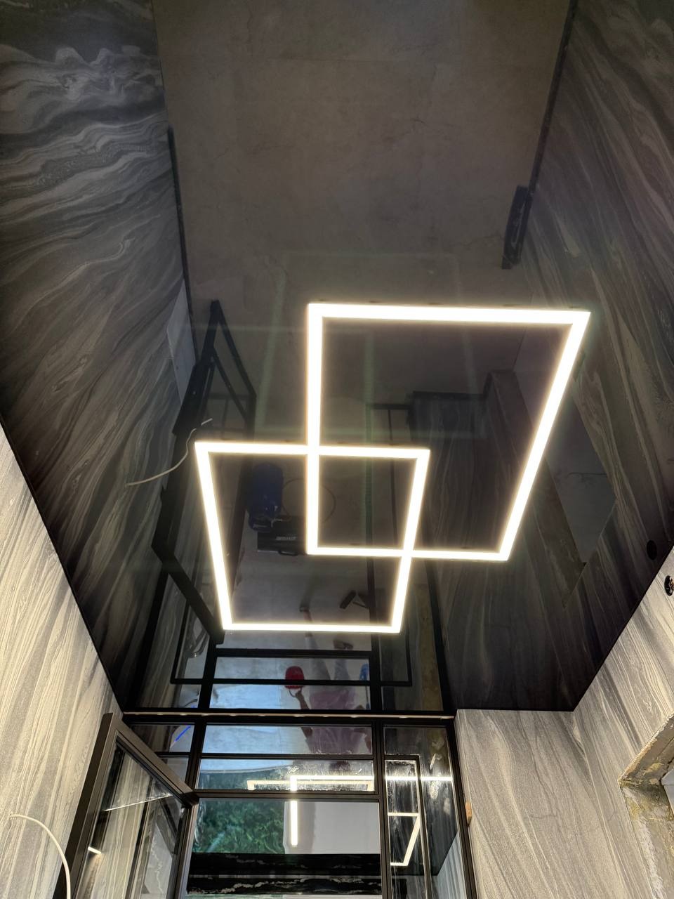 Błyszczący, ciemny sufit z efektem lustra, oświetlony dwoma kwadratowymi lampami LED. Widoczne odbicie pomieszczenia i okna. Ściany wykończone w szarym, marmurkowym wzorze.
