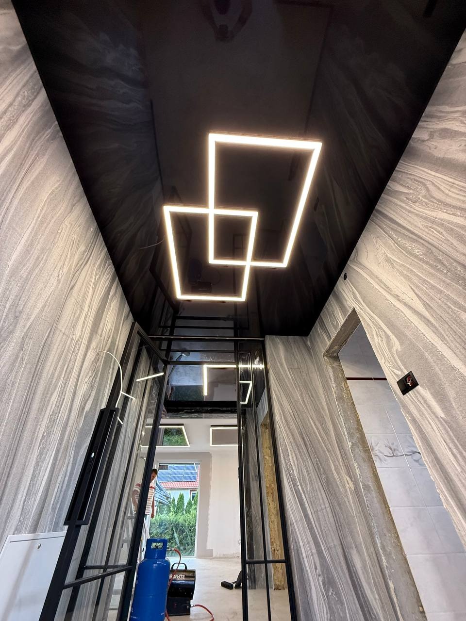 Błyszczący, czarny sufit z geometrycznym oświetleniem LED w kształcie kwadratów, w pomieszczeniu z szarymi ścianami o marmurkowym wzorze i czarną ramą drzwi.