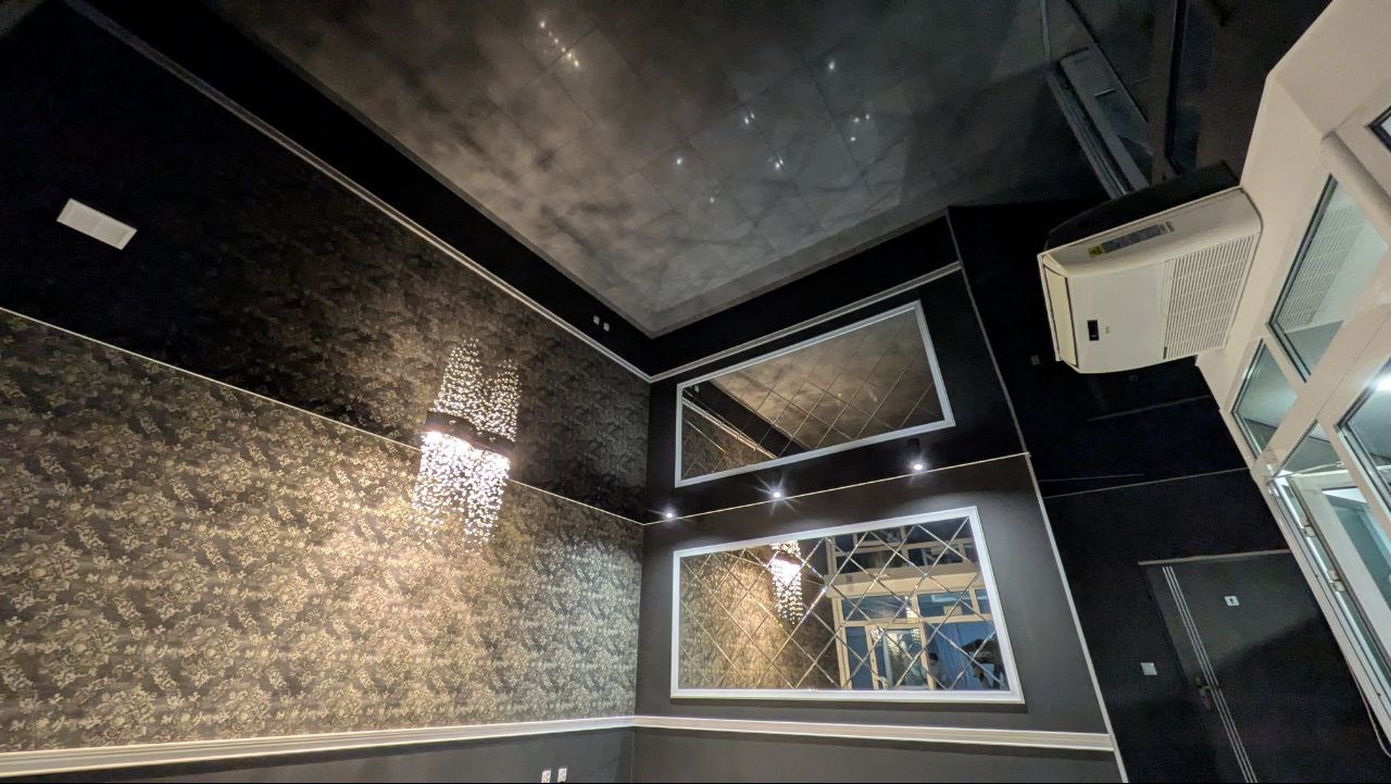 Eleganckie wnętrze z napinanym sufitem w odcieniach szarości, lustrzanymi panelami na ścianie i ozdobnym oświetleniem. Widoczny klimatyzator i drzwi.