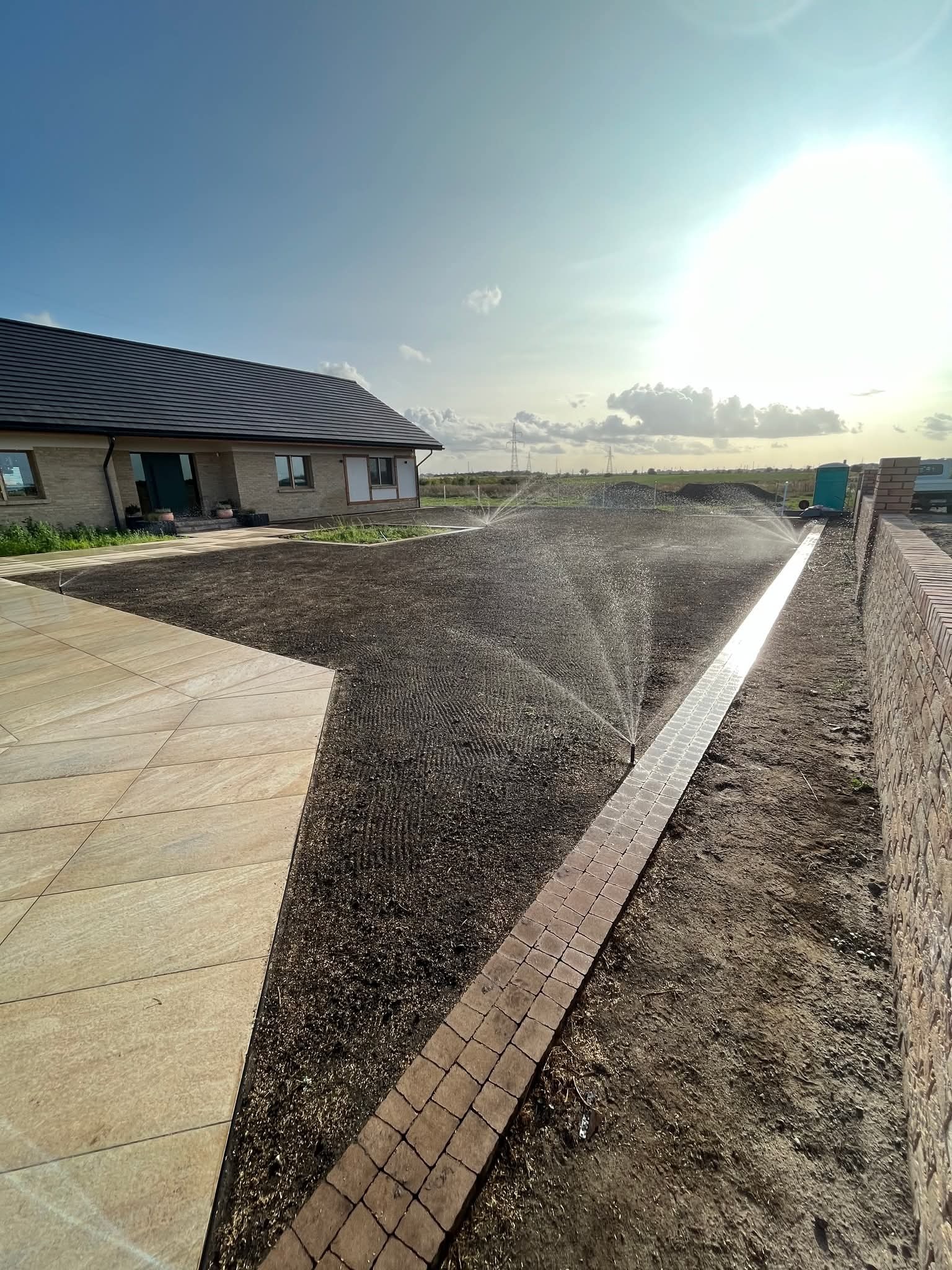 Nawadnianie nowo założonego trawnika z widocznymi zraszaczami i systemem rozprowadzającym wodę, w tle dom jednorodzinny i błękitne niebo.