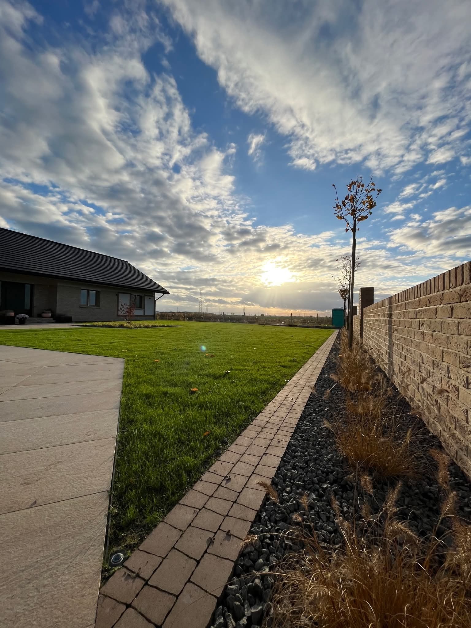 Nowoczesny ogród z równo przystrzyżonym trawnikiem, kamienną ścieżką i murem z cegły, oświetlony zachodzącym słońcem i dekoracyjnymi trawami.