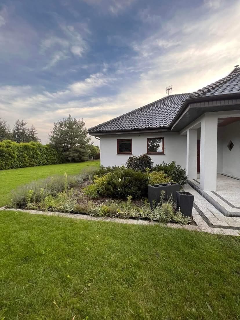 Ogród przy domu z rabatą kwiatową, trawnikiem i dwoma donicami przy wejściu. Trawa jest świeżo skoszona, a rabata starannie zagospodarowana. Widok na dom i fragment nieba.
