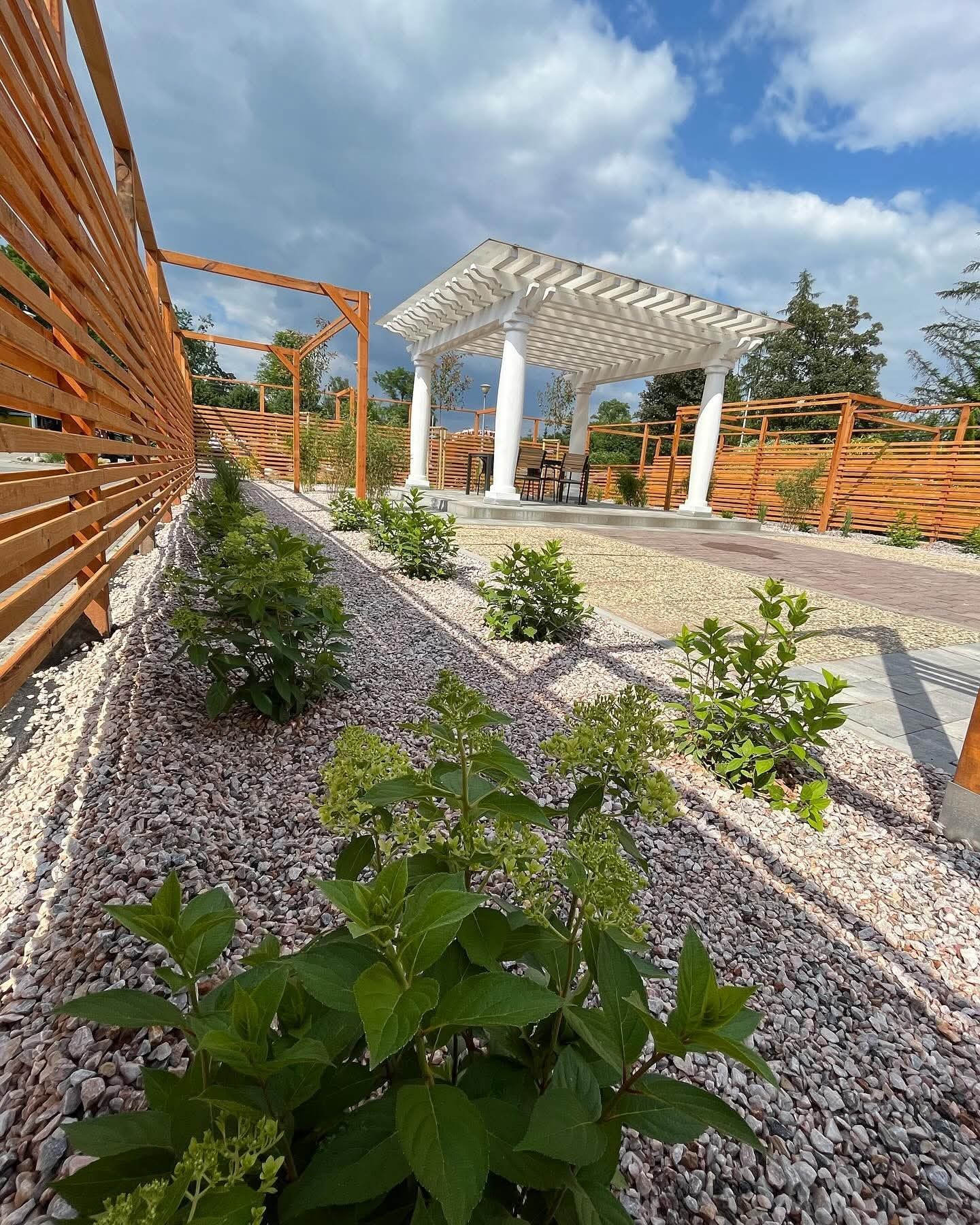 Ogród z białą altaną kolumnową, drewnianym ogrodzeniem i ścieżką z kamienia. Uwagę przykuwają zielone krzewy wzdłuż ścieżki i dekoracyjne kamyki.