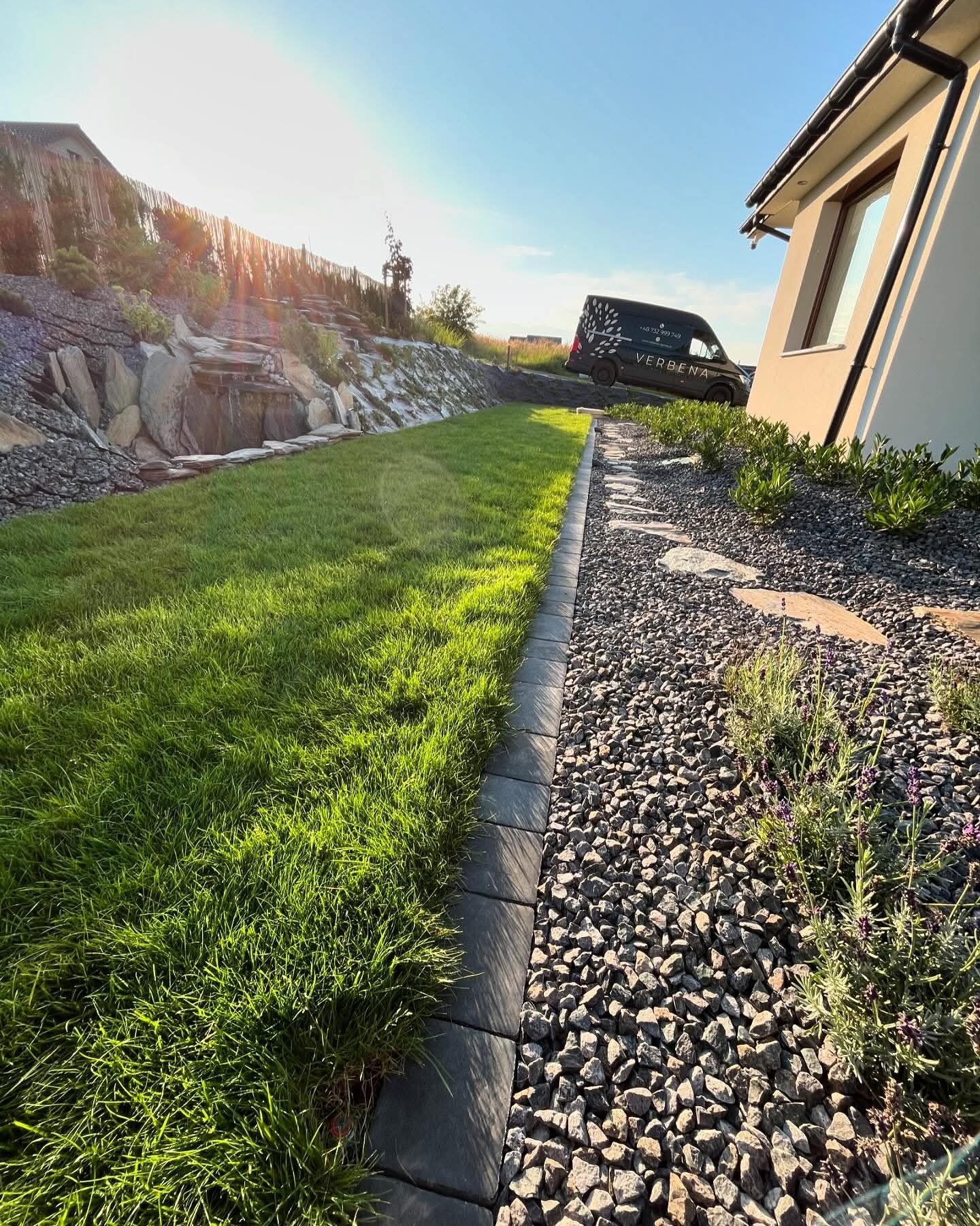 Ogród z równo przystrzyżonym trawnikiem, kamiennym obrzeżem i rabatą z kamyków oraz roślin. W tle widać wodospad i firmowy samochód.