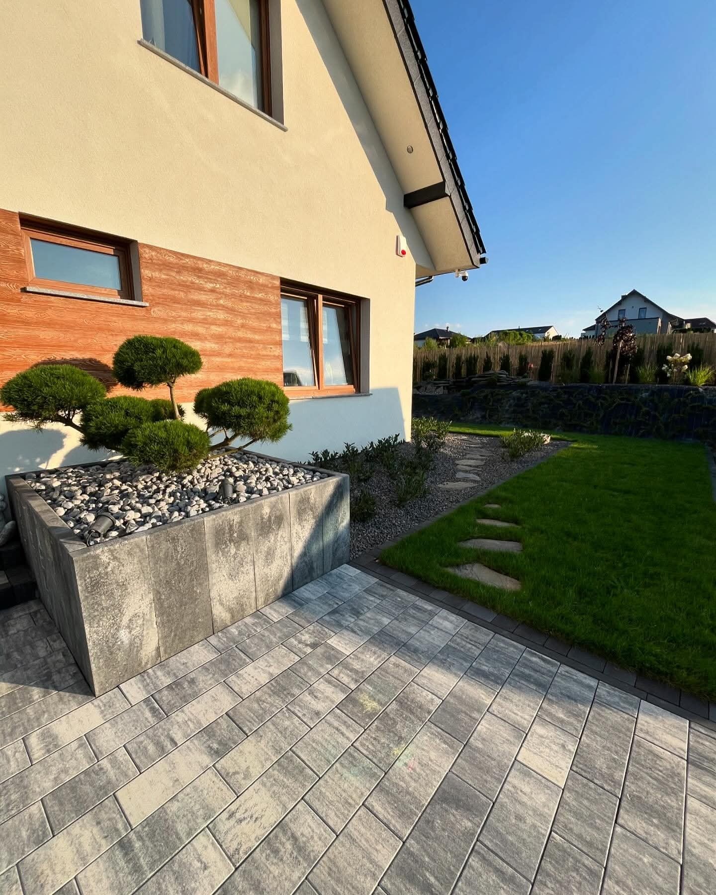 Nowoczesny ogród przydomowy z kostką brukową, betonową donicą z ozdobnymi drzewkami bonsai i kamieniami. Trawa, ścieżka z kamieni, budynek w tle.