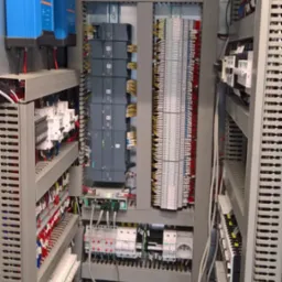 Rozbudowana szafa sterownicza z modułami Siemens, licznymi złączkami i okablowaniem, widok z bliska na skomplikowaną instalację elektryczną w szarej obudowie.