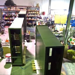 Volt Amper - Warsztat z pracownikami montującymi duże, metalowe szafy elektryczne. Widoczne narzędzia i komponenty, surowe oświetlenie, wysoki kąt widzenia z góry.