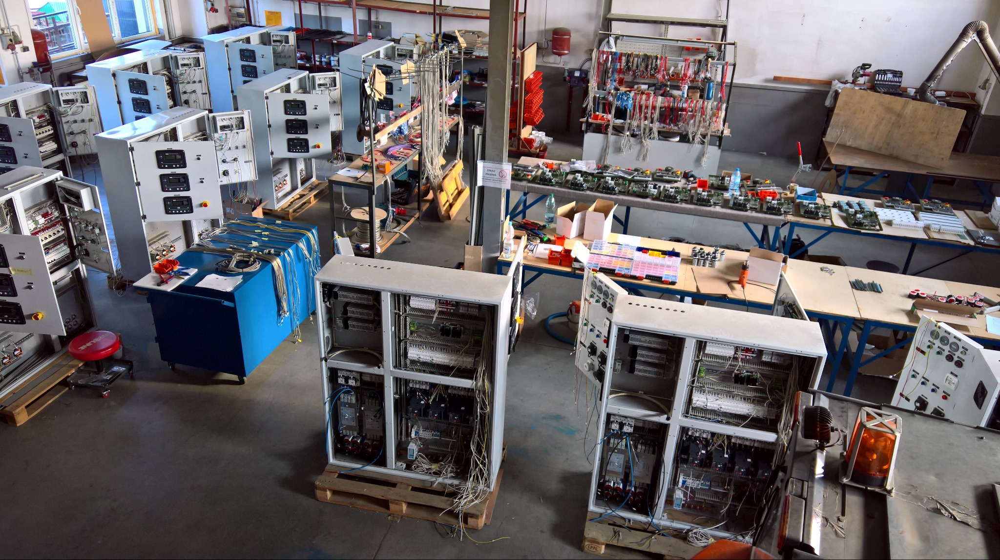 Warsztat z rozbudowanymi szafami sterowniczymi na paletach, licznymi narzędziami i elementami elektrycznymi na stołach. Widok z góry.