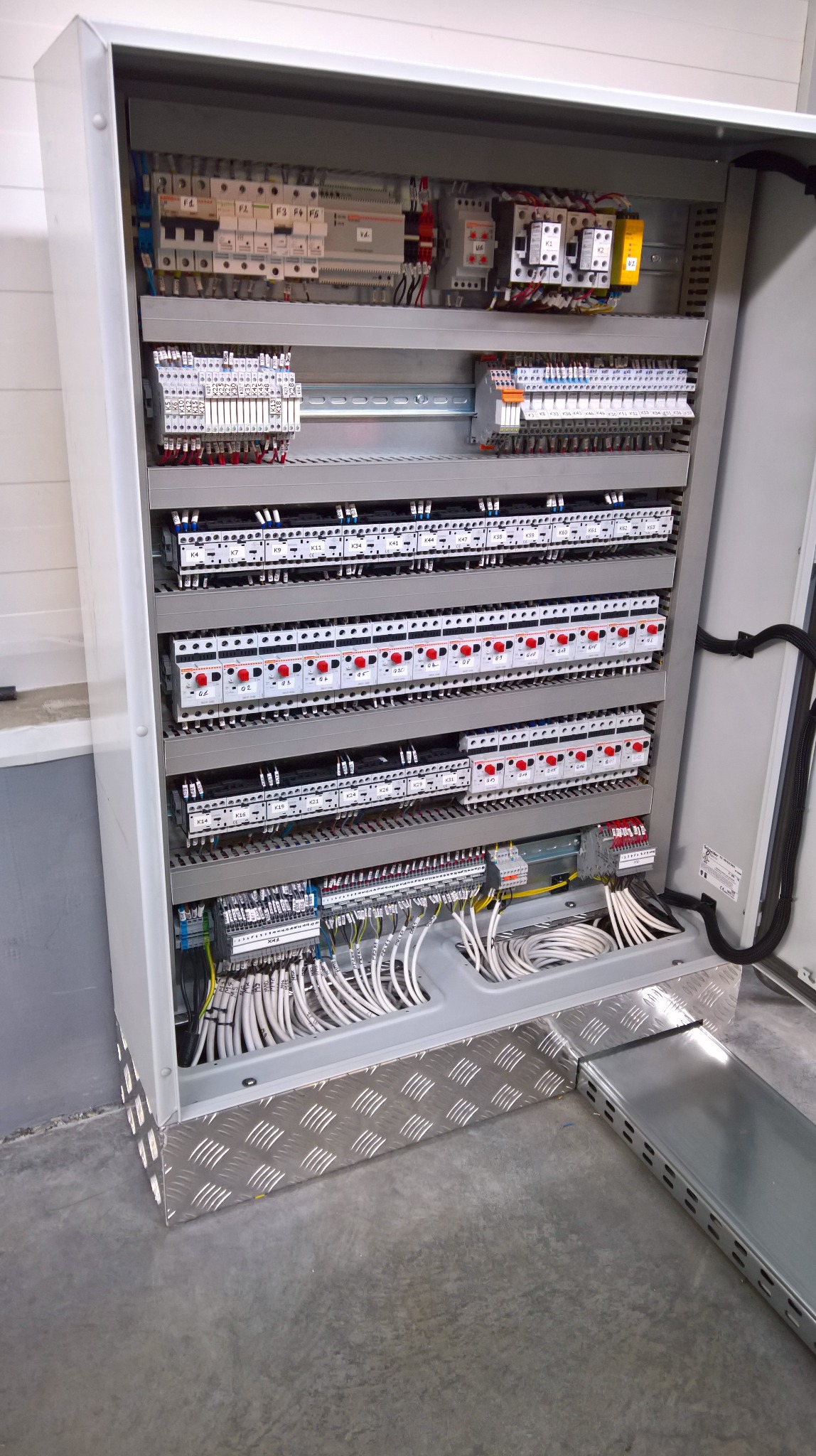 Otwarta, metalowa szafa sterownicza z widocznymi zabezpieczeniami, przekaźnikami i przewodami. Dolna część obudowy z blachy ryflowanej.