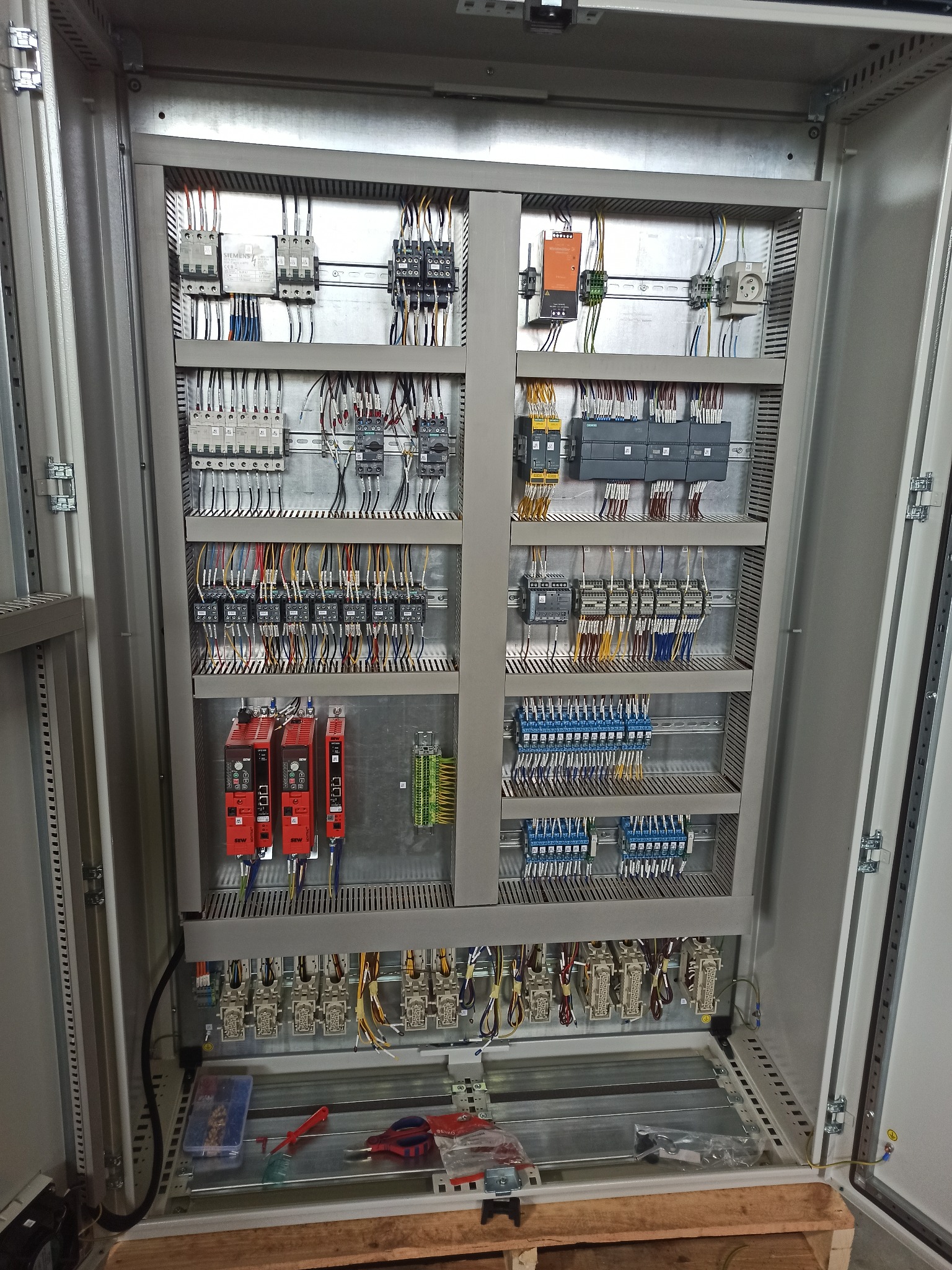 Rozdzielnica elektryczna w metalowej obudowie, widok z bliska na uporządkowane kable, moduły i elementy sterujące. Na dole widoczne narzędzia.