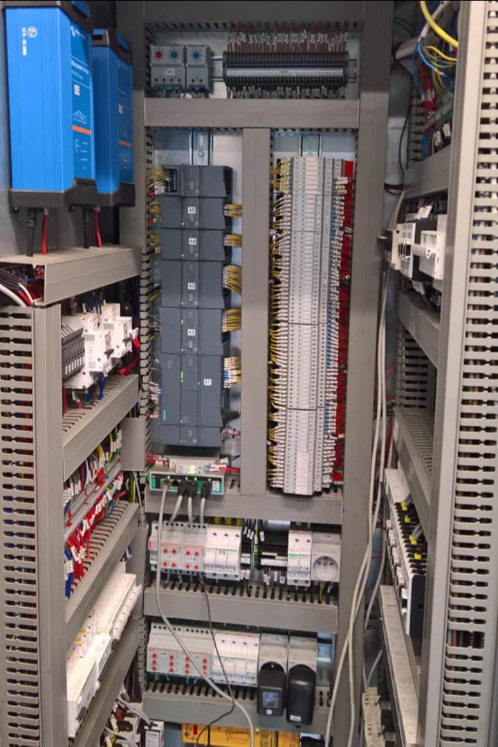 Rozbudowana szafa sterownicza z modułami Siemens, licznymi złączkami i okablowaniem, widok z bliska na skomplikowaną instalację elektryczną w szarej obudowie.