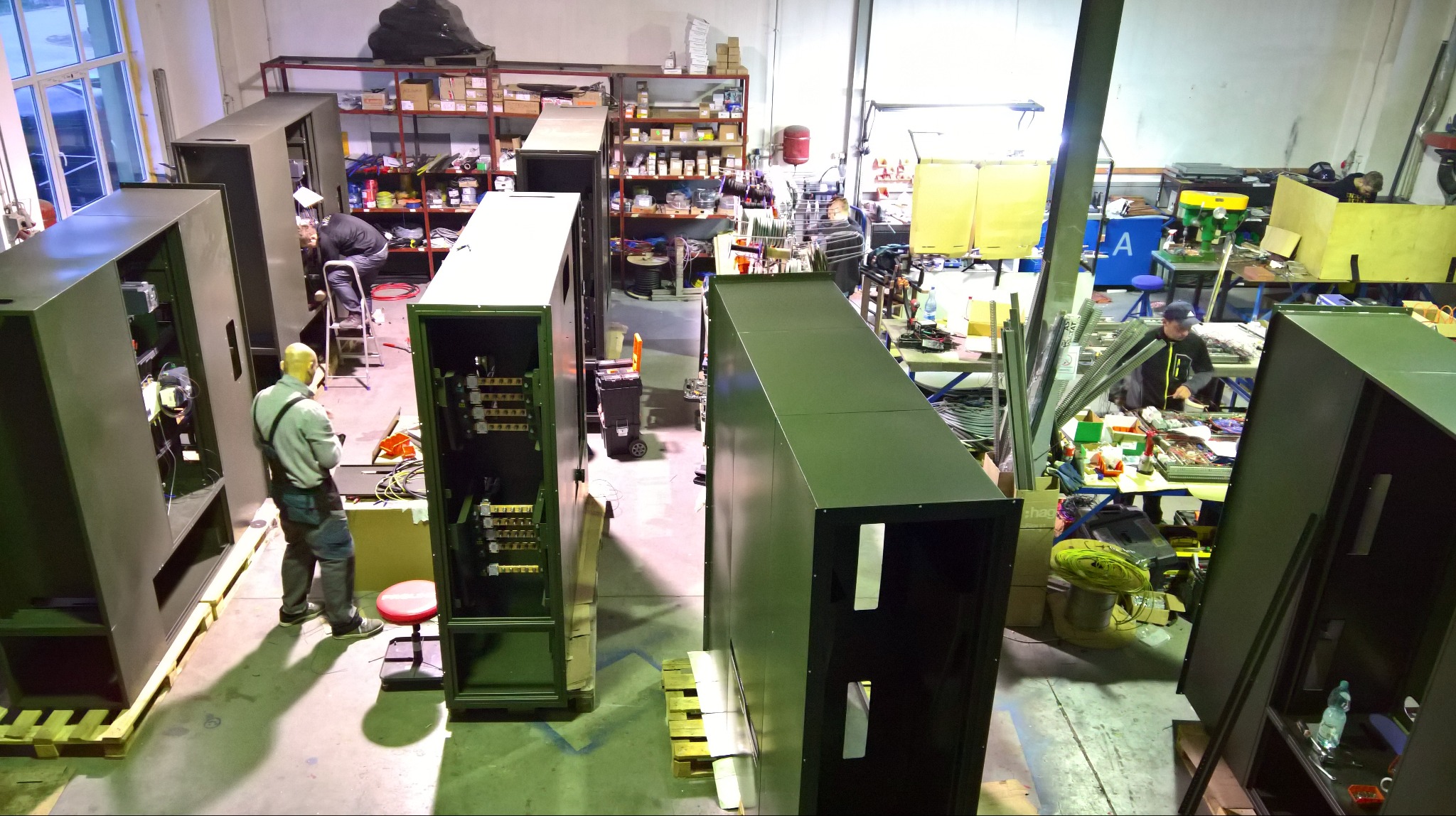 Warsztat z pracownikami montującymi duże, metalowe szafy elektryczne. Widoczne narzędzia i komponenty, surowe oświetlenie, wysoki kąt widzenia z góry.