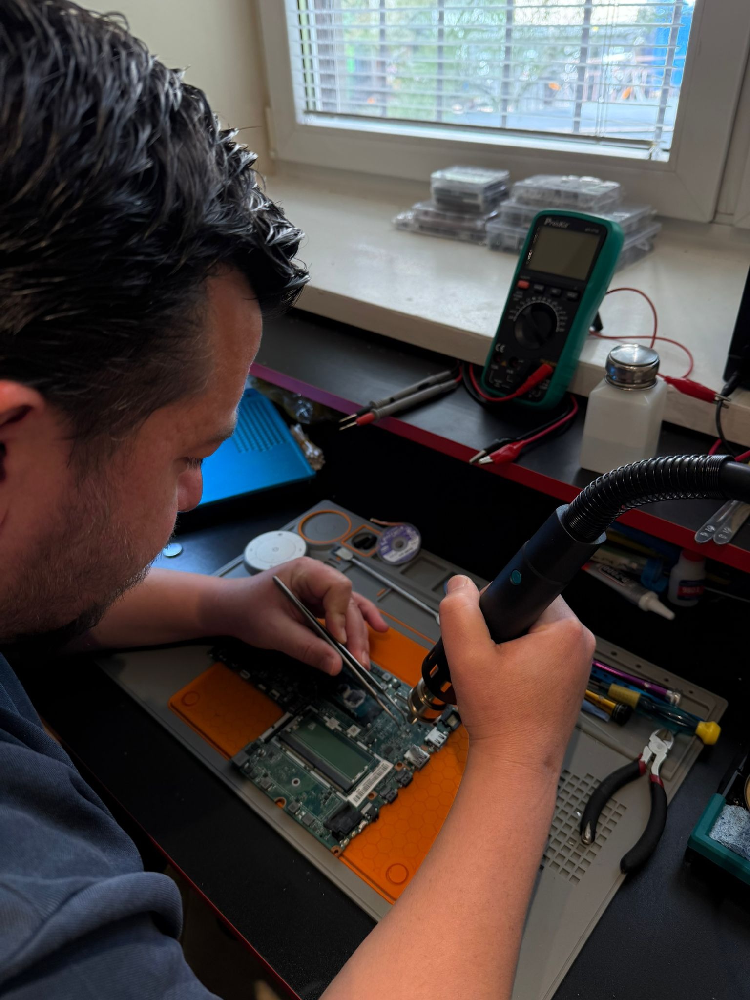 Serwisant precyzyjnie lutuje płytę główną laptopa przy użyciu pensety i hot air guna na macie ESD. W tle okno, multimetr i pojemniki na części.