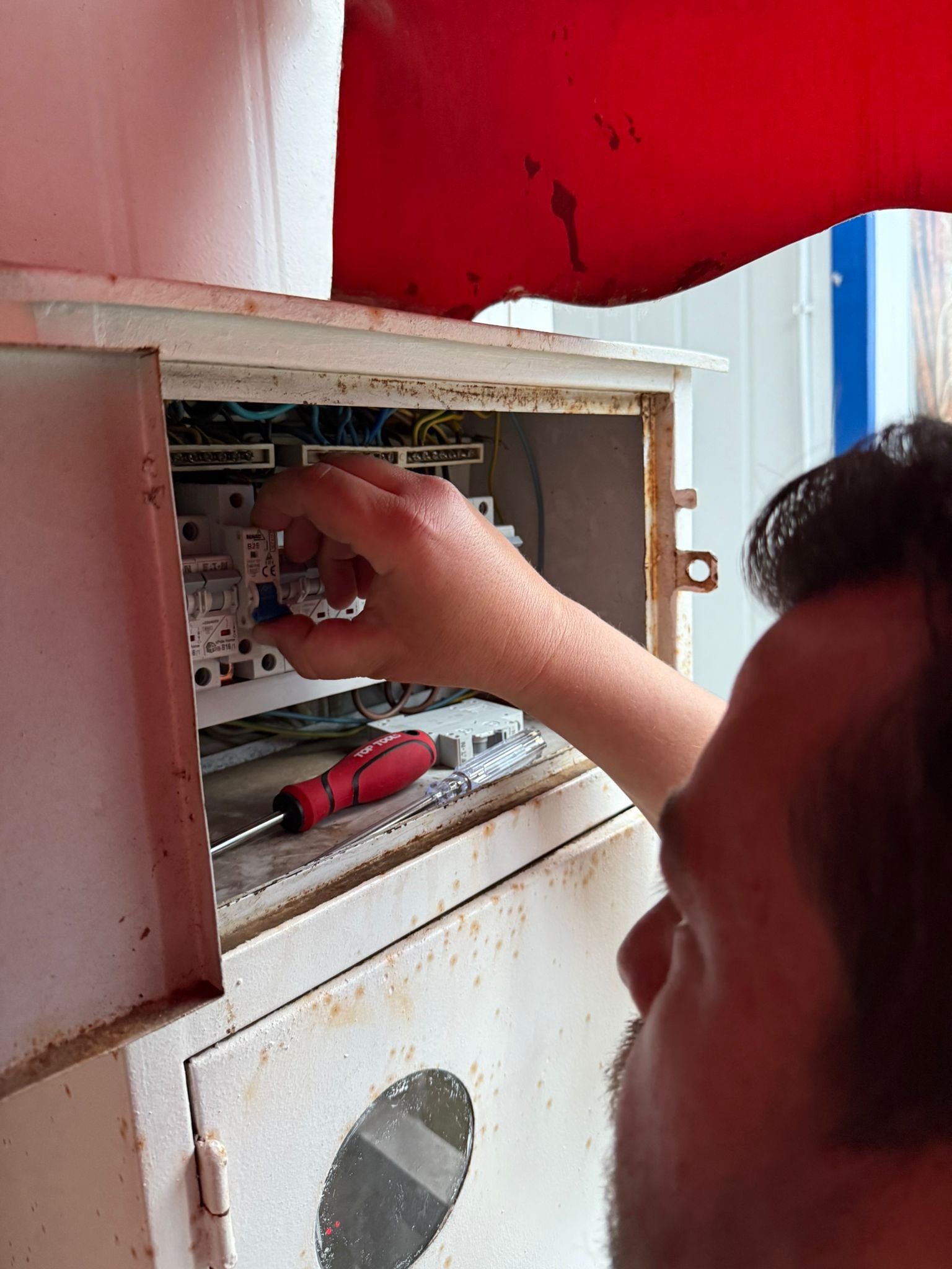 Elektryk w trakcie pracy nad instalacją elektryczną wewnątrz metalowej skrzynki, widoczne przewody, bezpieczniki i czerwony śrubokręt. Skrzynka nosi ślady korozji.