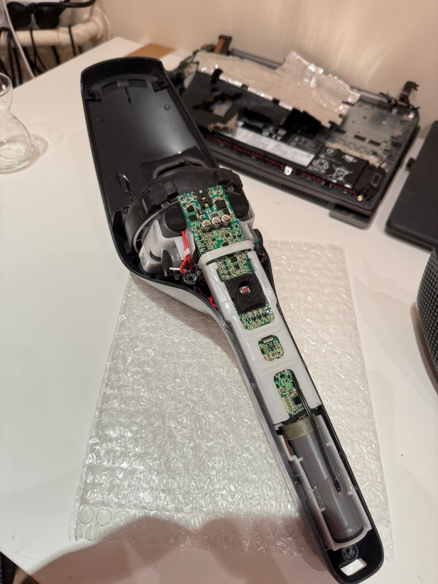 Otwarty, rozłożony odkurzacz bezprzewodowy na stole, ukazujący wewnętrzne komponenty elektroniczne i baterię. Obok widoczny rozmontowany laptop.