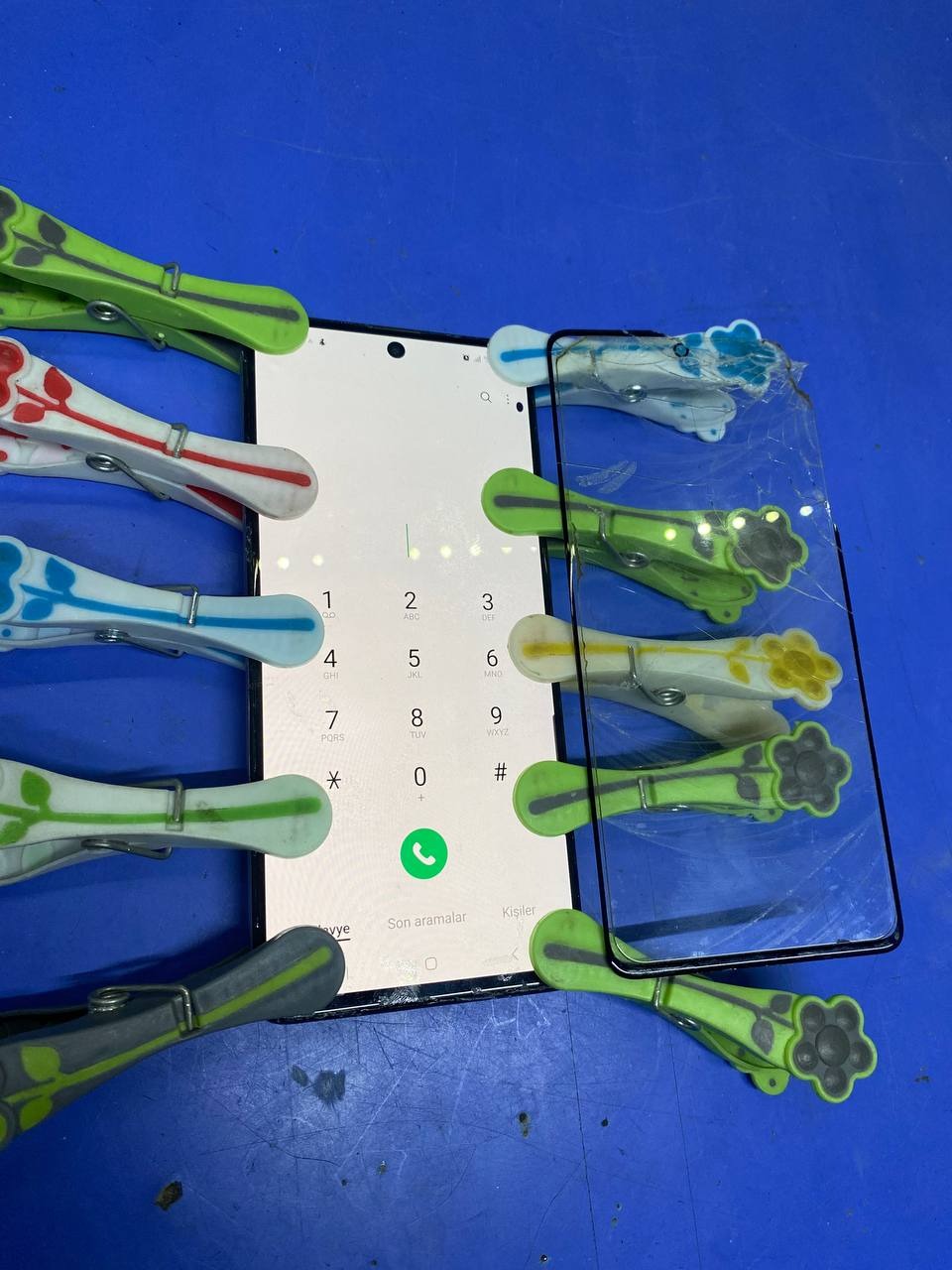 Smartfon z dialerem, dociskany klamerkami do przymocowania szkła ochronnego. Widoczne pęknięte szkło. Niebieskie tło.