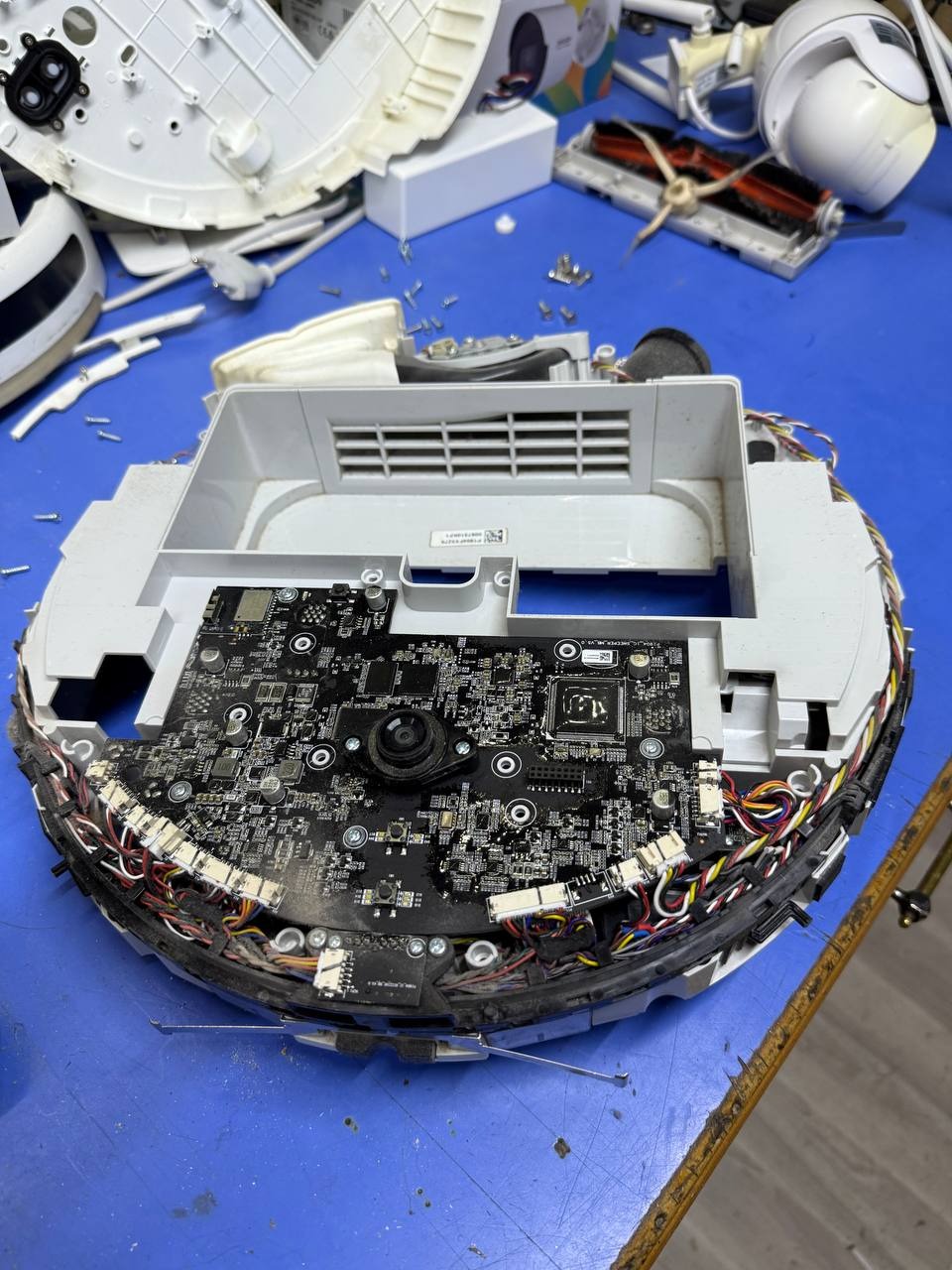 Rozłożony robot odkurzacz na niebieskim stole roboczym, ukazujący płytę główną, okablowanie i elementy obudowy w trakcie naprawy lub serwisu.