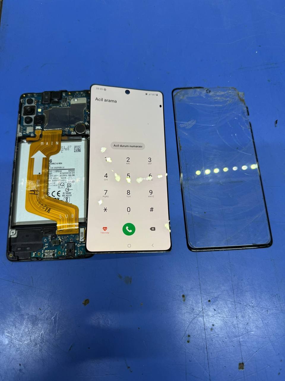 Rozłożony telefon Samsung: obudowa z elektroniką, ekran z klawiaturą numeryczną 'Acil arama' i uszkodzona szybka. Widok na niebieskim stole.