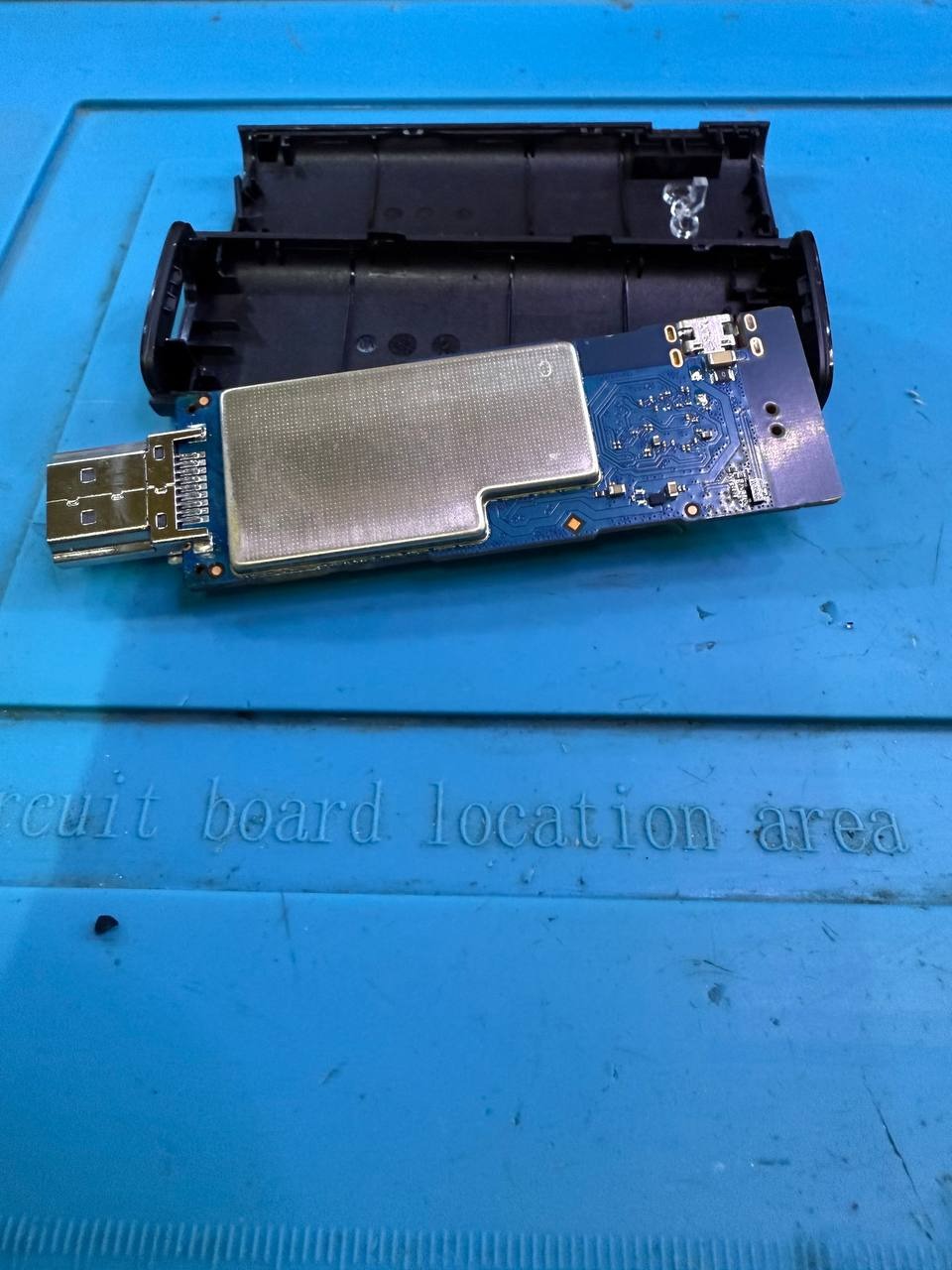 Otwarty moduł elektroniczny z USB i elementami na płytce, leżący na niebieskiej macie z napisem 'circuit board location area'.