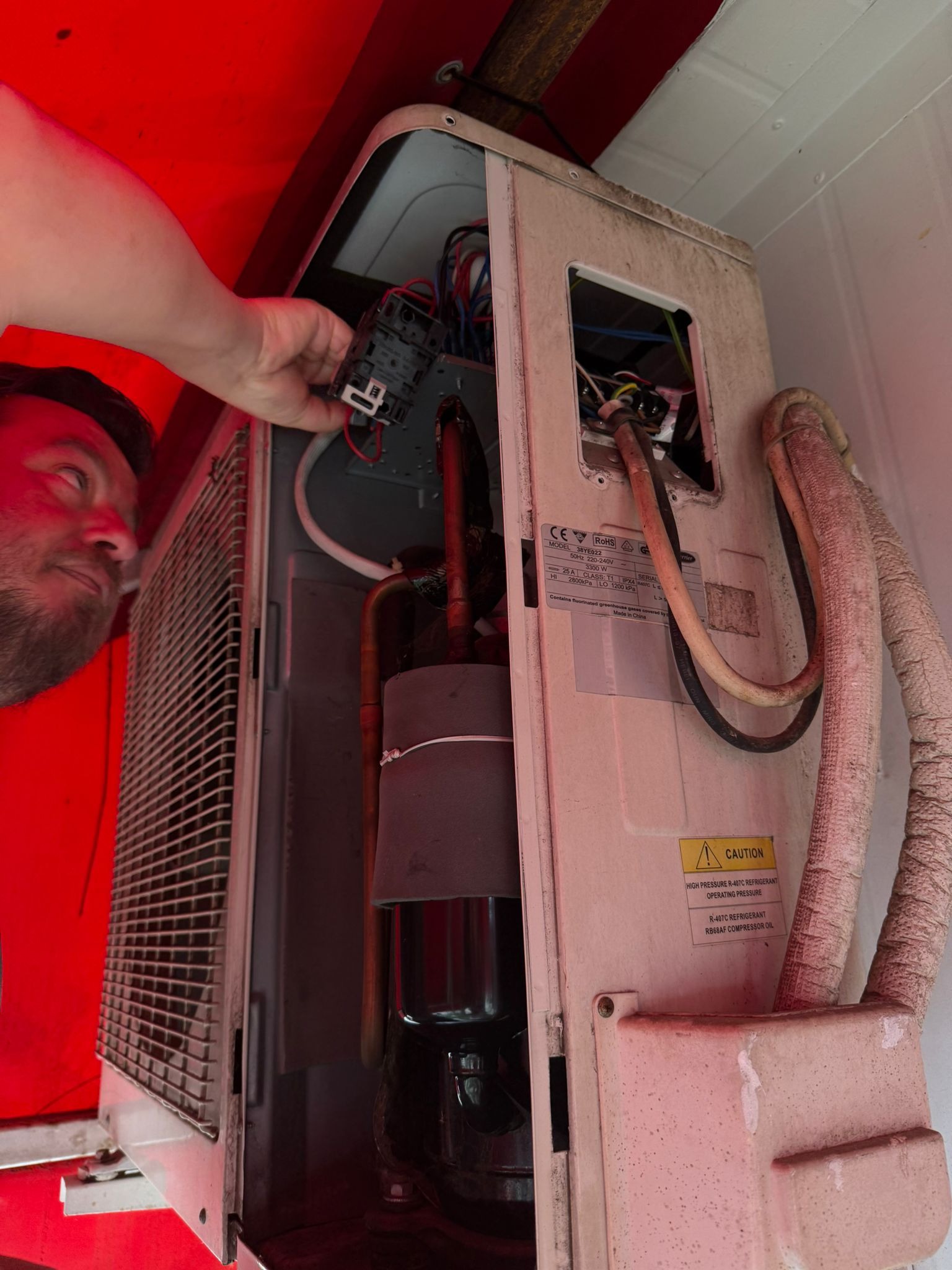 Elektryk podłącza moduł sterujący do klimatyzatora. Widoczne przewody, rury i panel sterowania. Urządzenie jest lekko zabrudzone, co sugeruje jego użytkowanie.