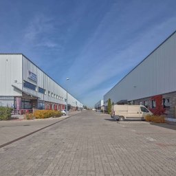 Kużnia Nieruchomości - Widok na ulicę z budynkami magazynowymi po obu stronach, zaparkowany van, kostka brukowa i błękitne niebo. Budynki z literą 'A' i nazwą firmy.