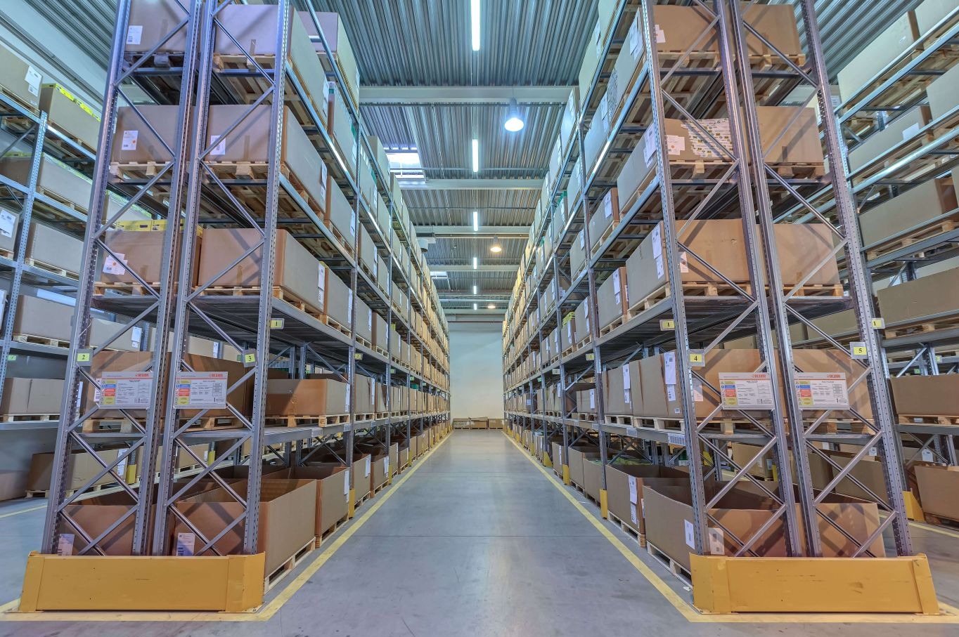 Wnętrze magazynu z wysokimi regałami wypełnionymi kartonami, oświetlone lampami sufitowymi. Widok w głąb korytarza między regałami, z żółtymi liniami na podłodze.
