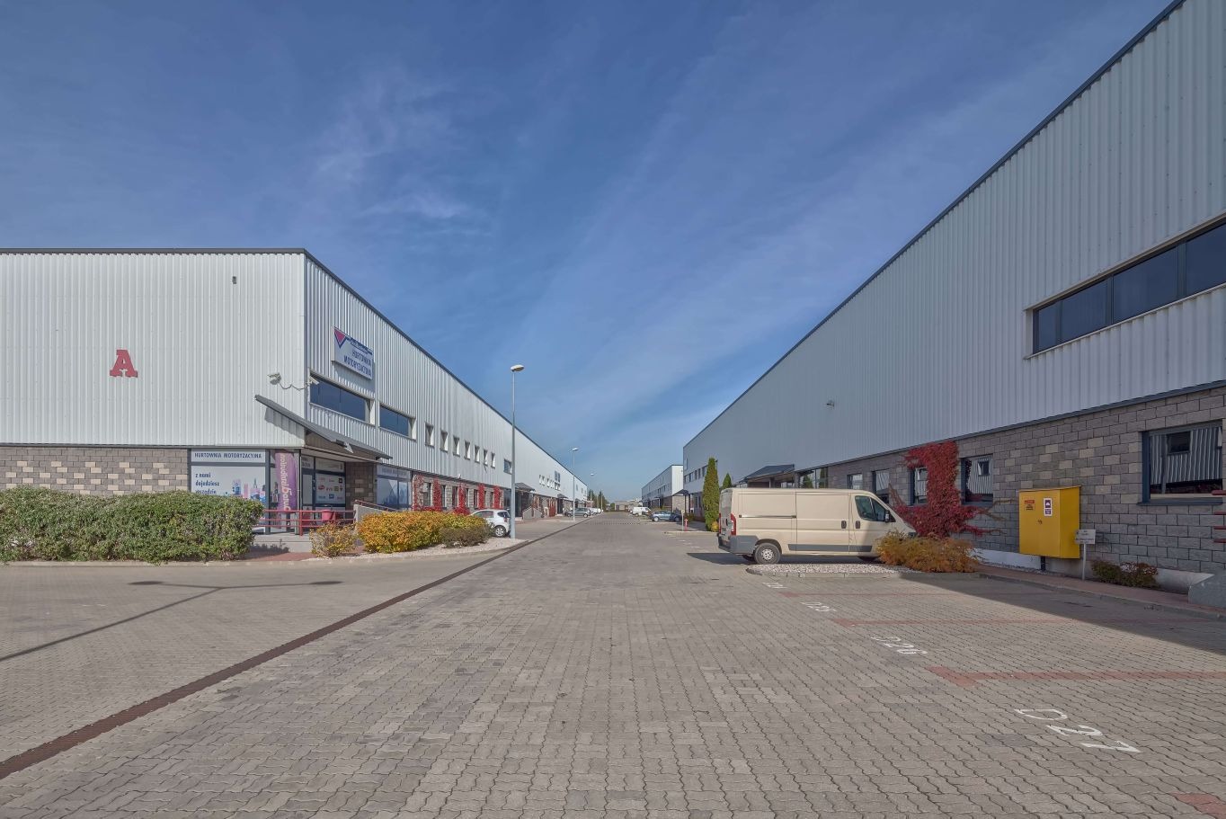 Widok na ulicę z budynkami magazynowymi po obu stronach, zaparkowany van, kostka brukowa i błękitne niebo. Budynki z literą 'A' i nazwą firmy.