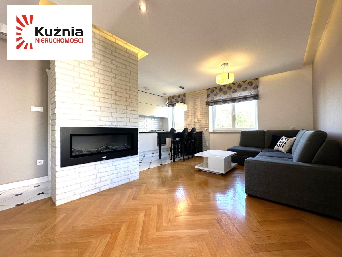 Jasne wnętrze mieszkania z parkietem w jodełkę, białym kominkiem i szarą sofą. Widok na kuchnię i okno z roletą rzymską. Logo agencji nieruchomości w rogu.