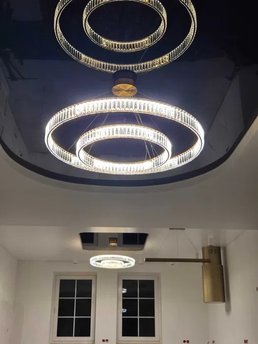 Błyszczące, okrągłe lampy wiszące z kryształkami, zamontowane w błyszczącym, czarnym suficie. Widok na wnętrze z białymi ścianami i oknami.