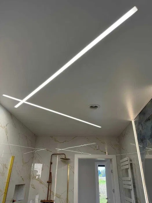 Nowoczesna łazienka z marmurowymi ścianami i innowacyjnym oświetleniem sufitowym w postaci krzyżujących się, liniowych lamp LED. Widok na prysznic i grzejnik.