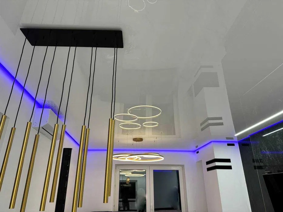 Błyszczący sufit napinany z nowoczesnym oświetleniem: podłużna lampa z złotymi elementami i okrągłe lampy. Niebieskie światło LED podkreśla krawędzie sufitu i ściany.