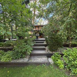 Przemysław gaca - Ogród z drewnianymi schodami prowadzącymi do domu, otoczony bujną zielenią i kamieniami. Roślinność różnorodna, z elementami dekoracyjnymi. Ujęcie z dołu, w kierunku domu.