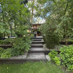 Ogród z drewnianymi schodami prowadzącymi do domu, otoczony bujną zielenią i kamieniami. Roślinność różnorodna, z elementami dekoracyjnymi. Ujęcie z dołu, w kierunku domu.