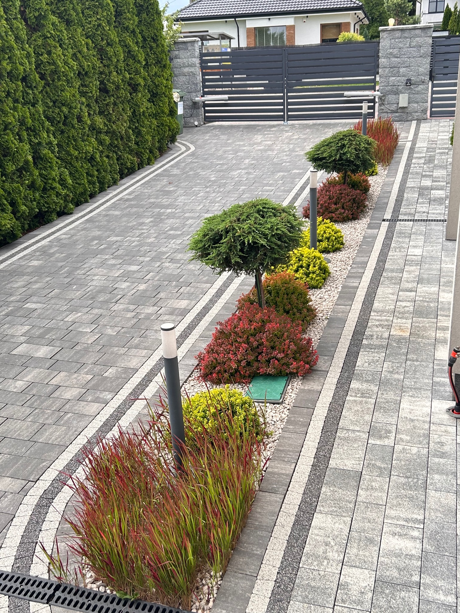 Ogród z równo przyciętymi krzewami i drzewami ozdobnymi wzdłuż ścieżki z kostki brukowej, Nowy Dwór Mazowiecki. Oświetlenie ogrodowe.