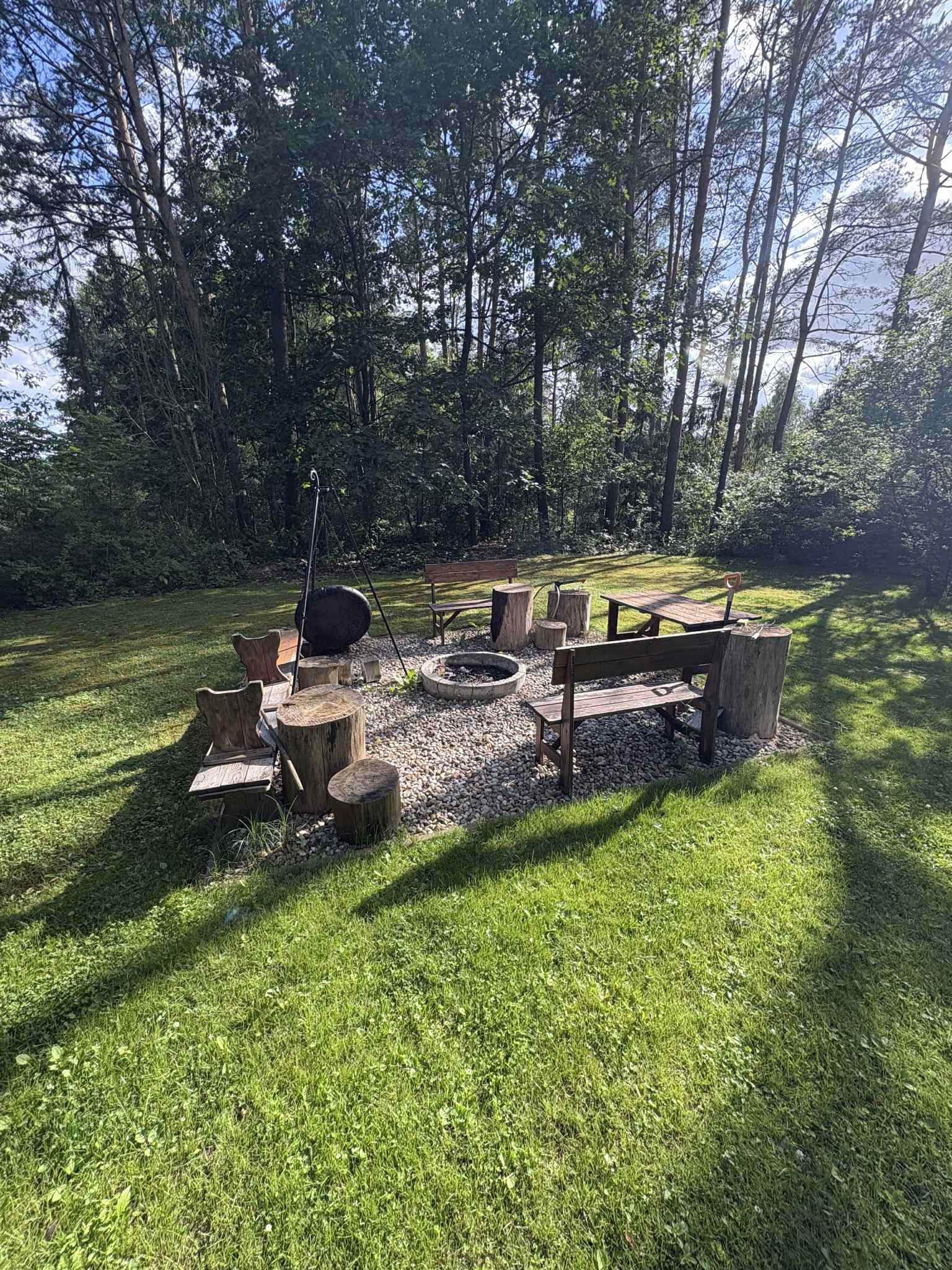 Ognisko otoczone drewnianymi siedziskami i stołem, wyłożone kamieniami, z kotłem na trójnogu, na tle zielonego trawnika i drzew.