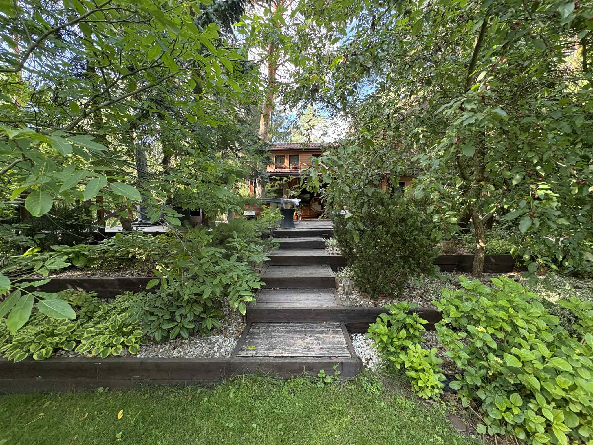Ogród z drewnianymi schodami prowadzącymi do domu, otoczony bujną zielenią i kamieniami. Roślinność różnorodna, z elementami dekoracyjnymi. Ujęcie z dołu, w kierunku domu.