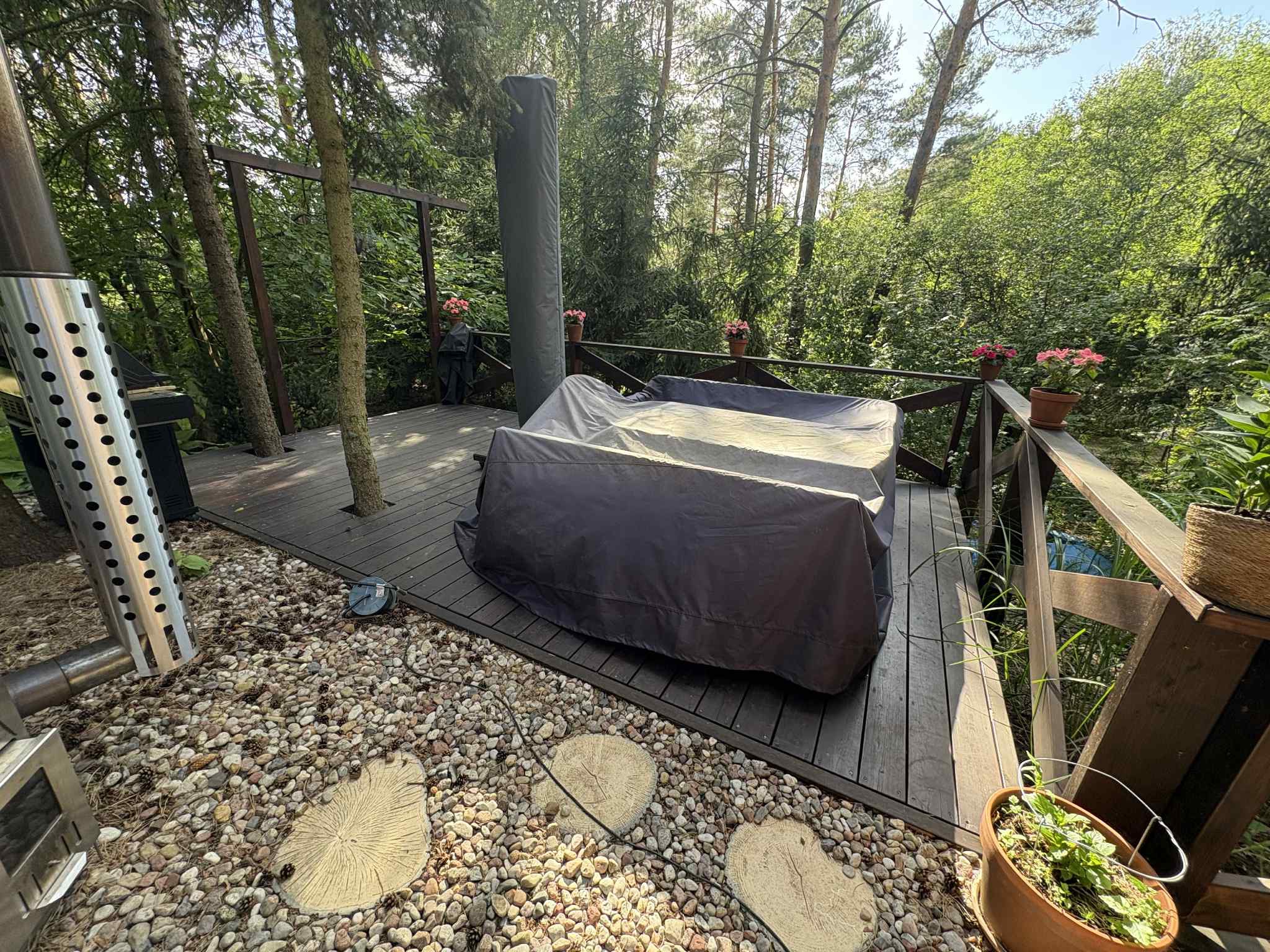 Drewniany taras z jacuzzi pod pokrowcem, otoczony zielenią. Dekoracyjne kamienie i donice z kwiatami dodają uroku. Widoczny piec do jacuzzi.
