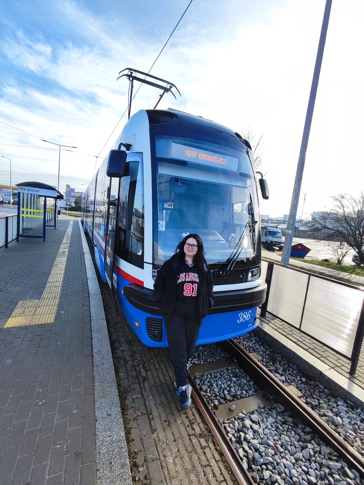 Dziewczyna na tle tramwaju MZA Bydgoszcz na przystanku Wyżyny w słoneczny dzień. Tramwaj o numerze 386, biało-niebieski z czerwonym paskiem.