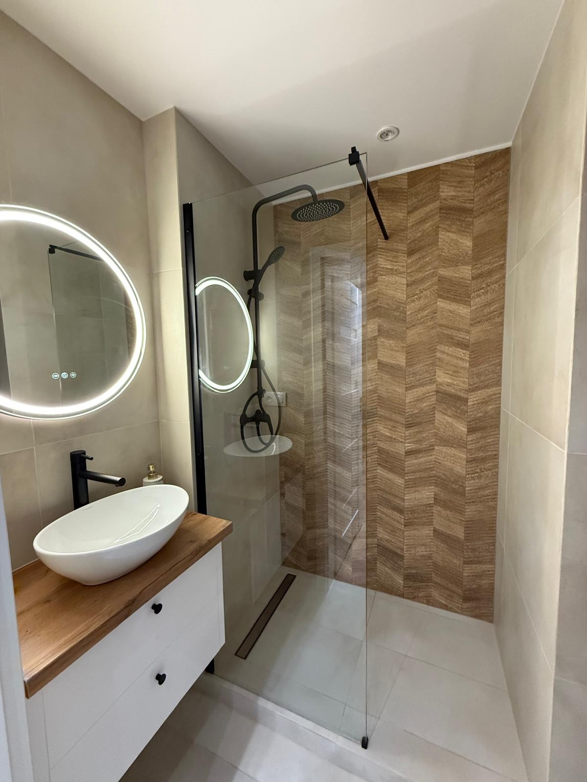 Nowoczesna łazienka z kabiną typu walk-in, umywalką nablatową na drewnianym blacie i okrągłym lustrem z podświetleniem LED. Ściana kabiny wykończona płytkami w jodełkę.